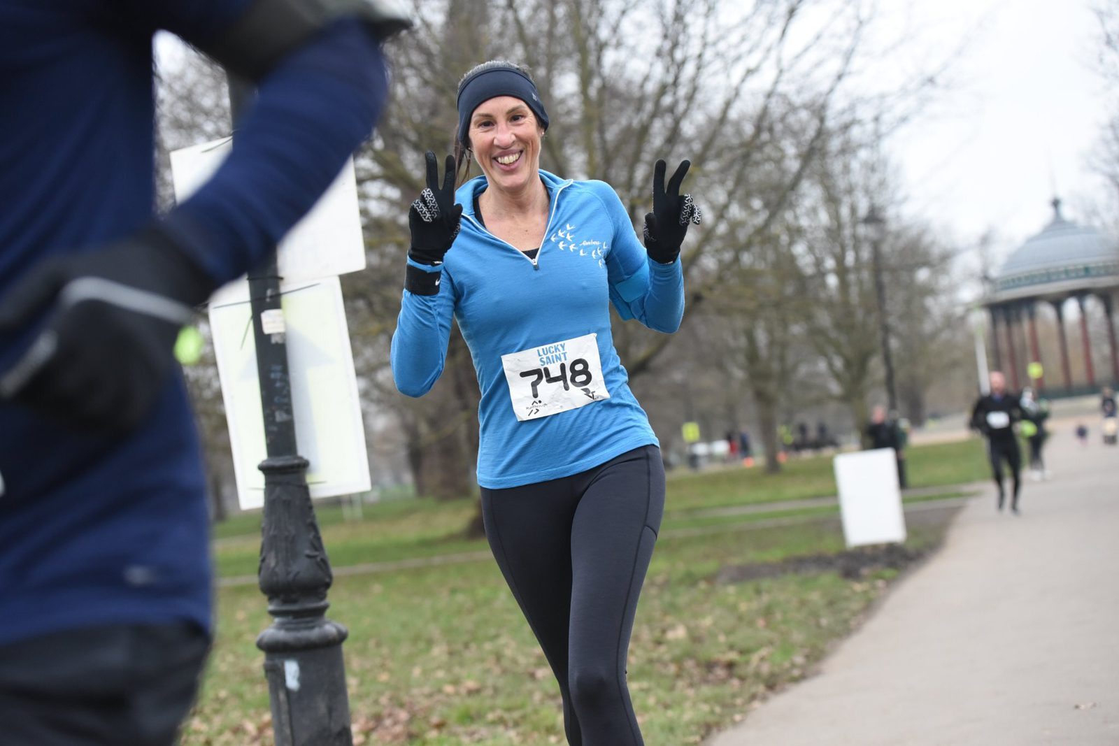 A woman in a blue long-sleeve shirt and black leggings runs in a park, smiling and showing a peace sign with both hands. She wears a race bib numbered 748. Other runners and bare trees are visible in the background.