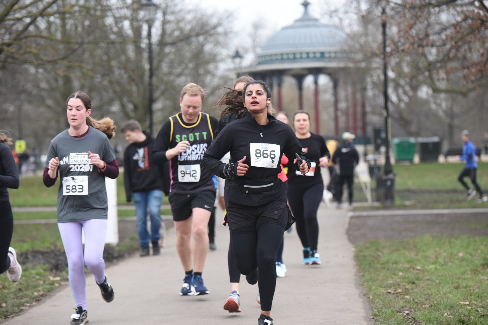 Runners participating in a race on a park pathway, with trees and a gazebo in the background. They wear numbered bibs and athletic clothing. The environment is overcast, and spectators can be seen in the distance.