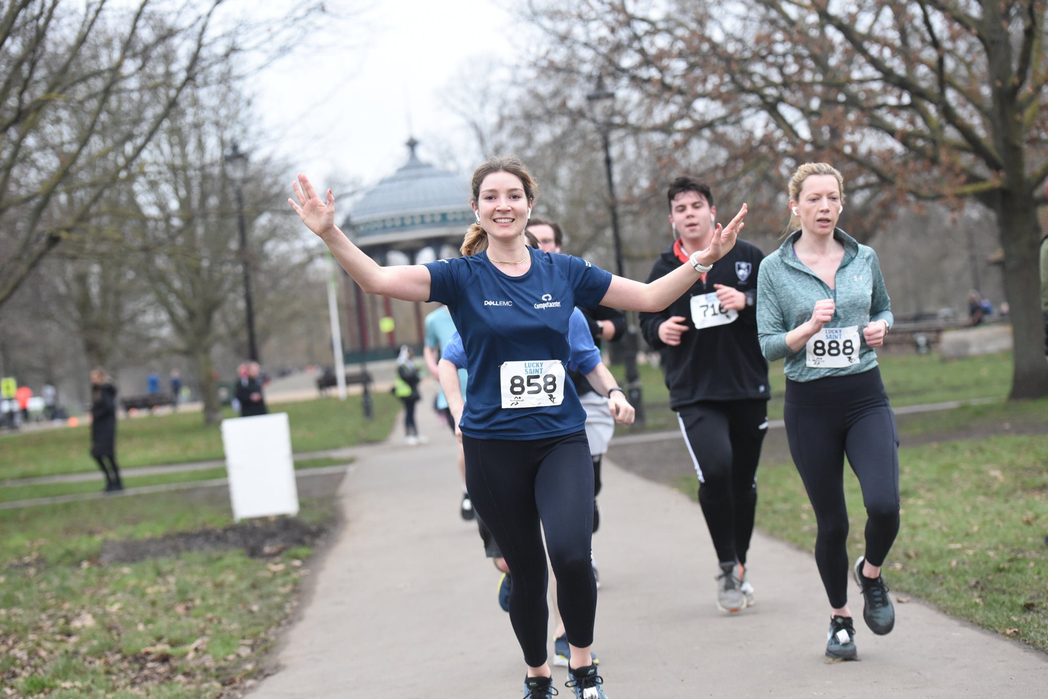 A group of people running on a park path. The woman in front, wearing a blue shirt and black leggings, is smiling and raising her arms. Other runners follow behind, with trees and a pavilion visible in the background.