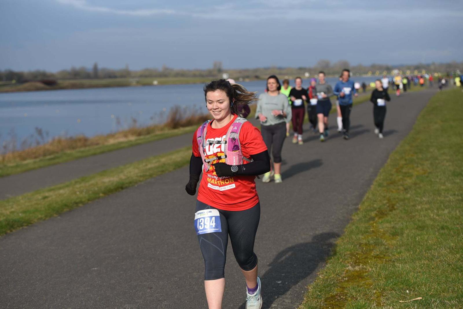 A woman in a red marathon shirt and black leggings runs on a paved path beside a body of water, leading a group of runners. It's a clear day and the landscape is green and open.