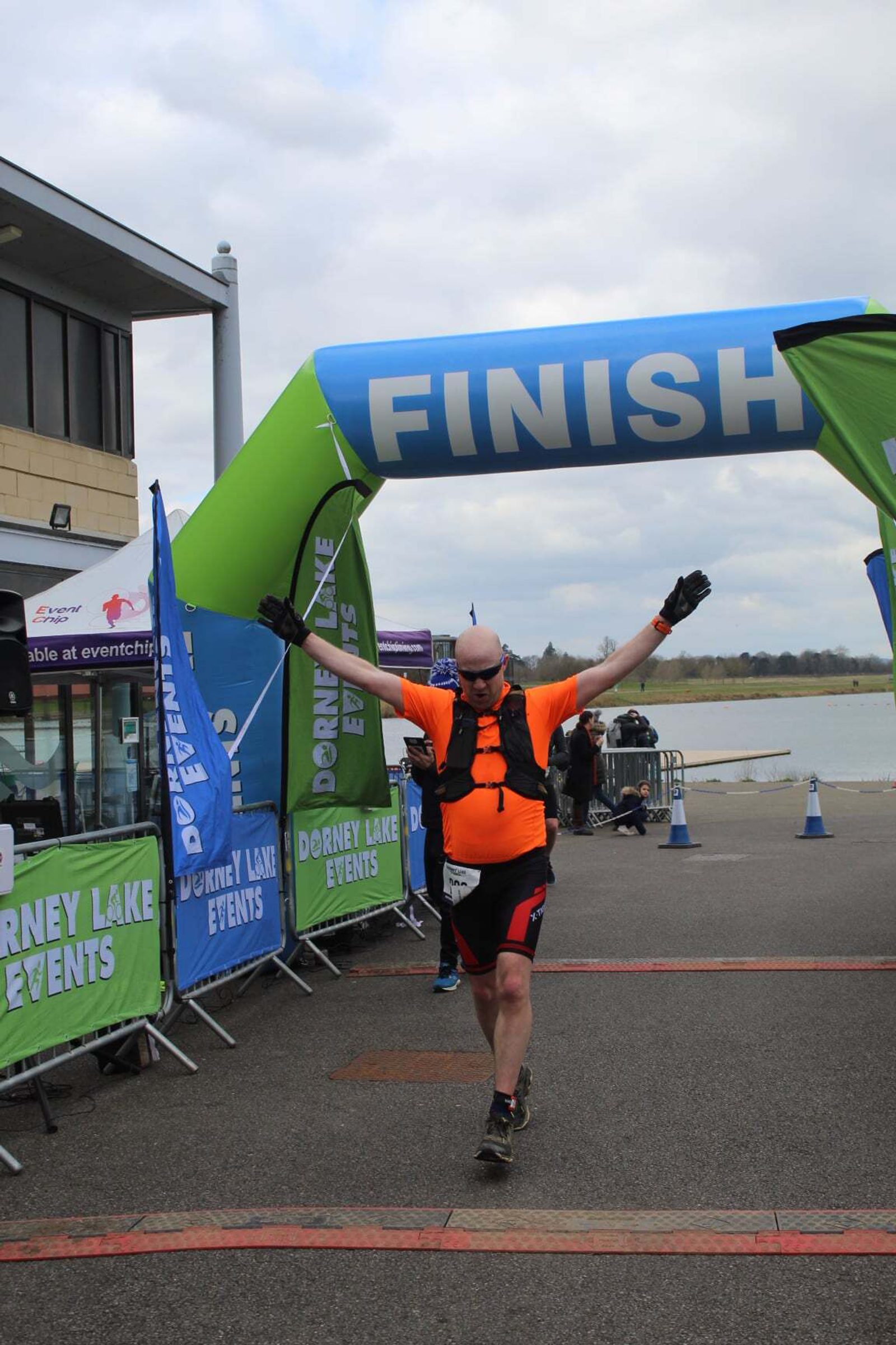 A runner wearing an orange shirt crosses the finish line at an outdoor event near a lake. The banners read "Dorney Lake Events." The sky is cloudy and there are cones and a building in the background.