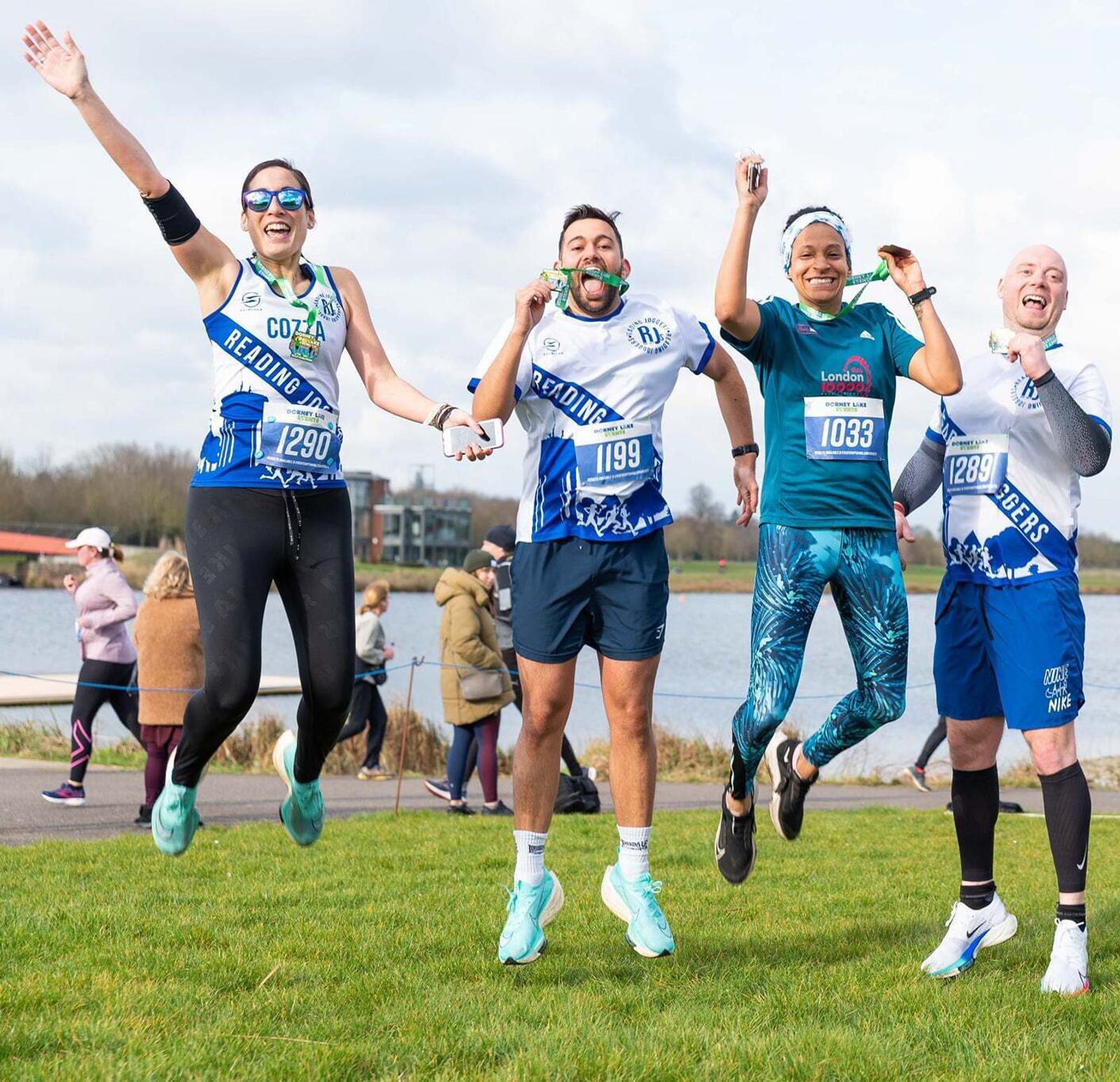 Five runners are celebrating and jumping in the air by a lake. They are wearing race bibs and sports attire. The sky is partly cloudy, and other people are visible in the background.