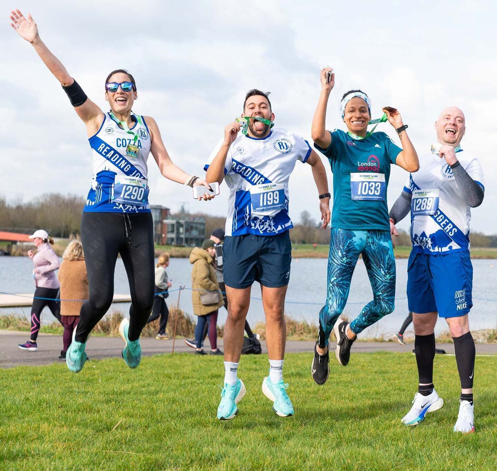 Five people in athletic wear joyfully jump in the air with medals around their necks, standing on grass near a body of water. They appear to have just completed a race, celebrating with smiles and energetic poses.