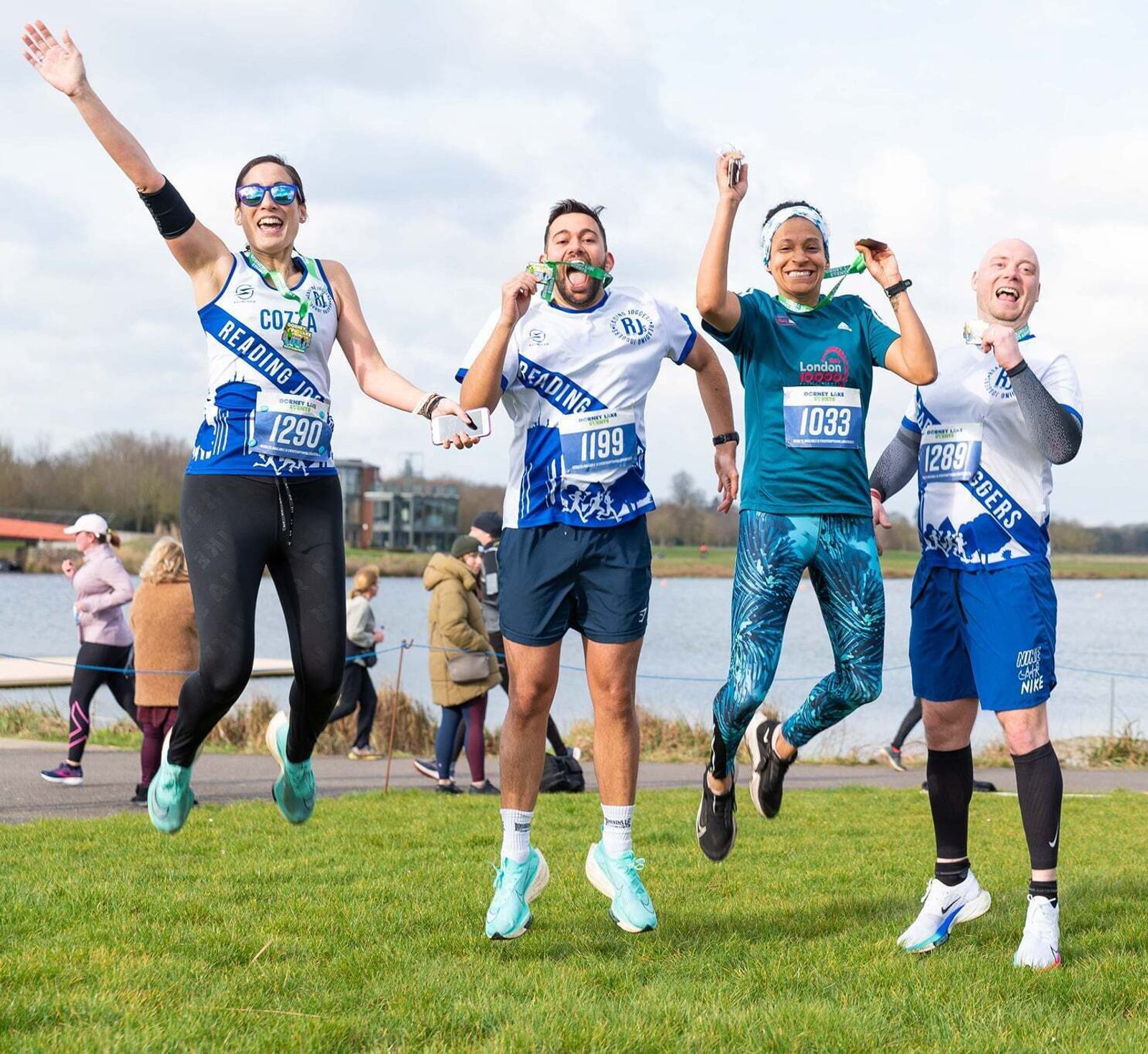 Five runners in colorful athletic gear jump in the air celebrating after a race near a body of water. They are smiling and appear energetic. Onlookers and a cloudy sky are visible in the background.