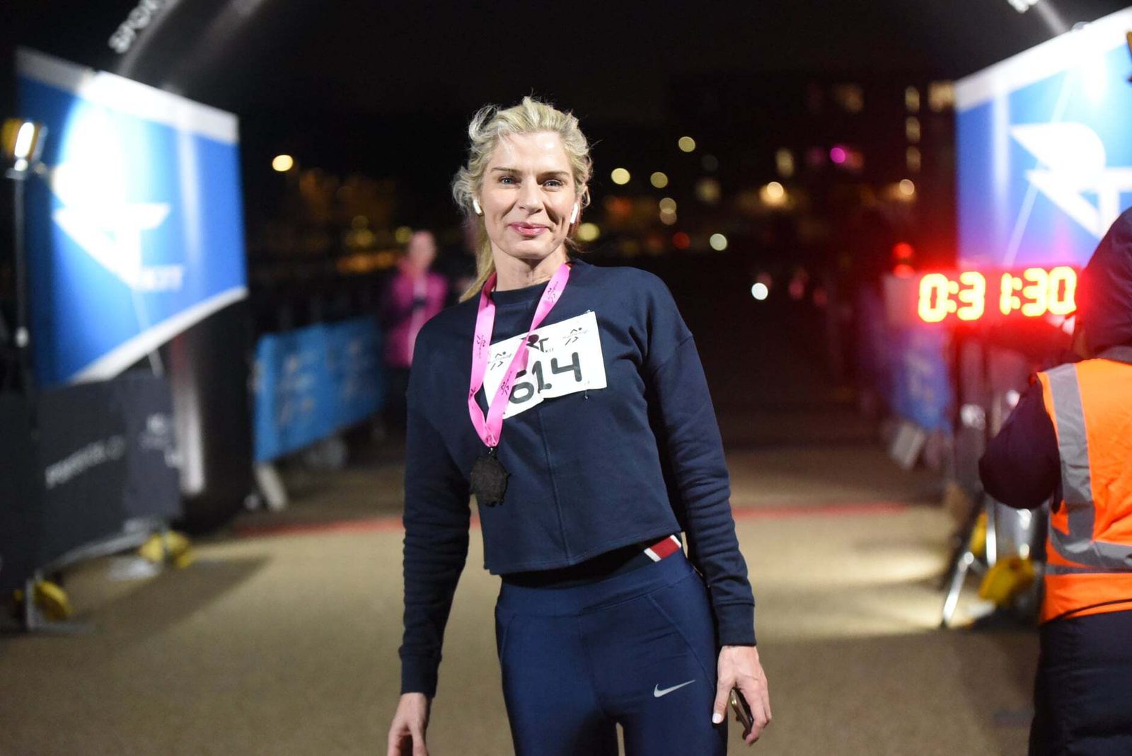 A person stands confidently at a race finish line at night, wearing a numbered race bib and a medal. The illuminated clock in the background displays the time 03:13:04.