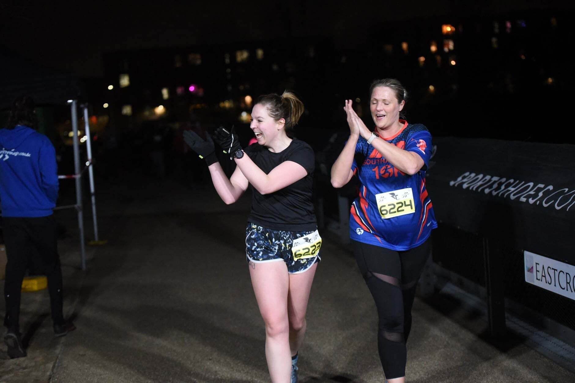 Two women are joyfully clapping while walking at night in a running event. Both wear race bibs; one in a black outfit, the other in a dark blue shirt with colorful details. Background includes blurred lights and a partially visible event banner.