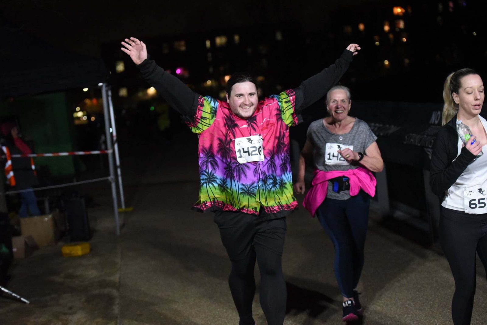 A person wearing a colorful tie-dye shirt raises their arms triumphantly while running in a night race. Two other participants run nearby, all wearing race numbers. The background shows a dimly lit urban scene.