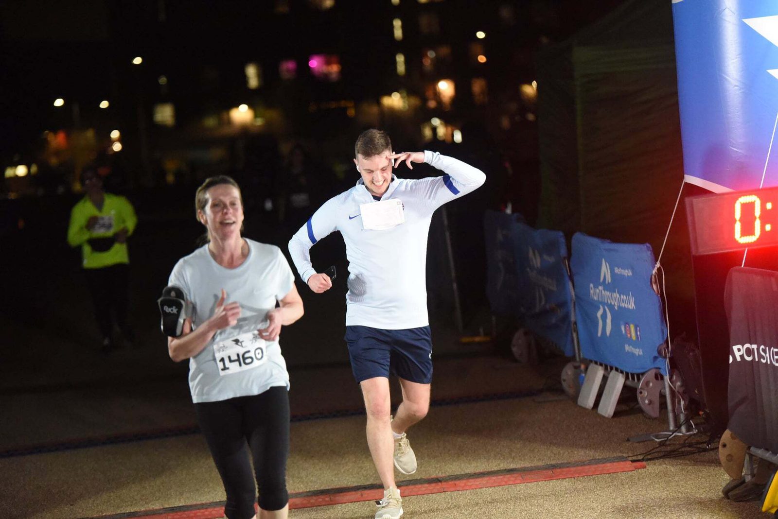 Two runners crossing a finish line at night. The woman wears a white shirt with the number 1460, smiling as she finishes. The man beside her touches his ear while running. A large digital clock and blurred lights are visible in the background.