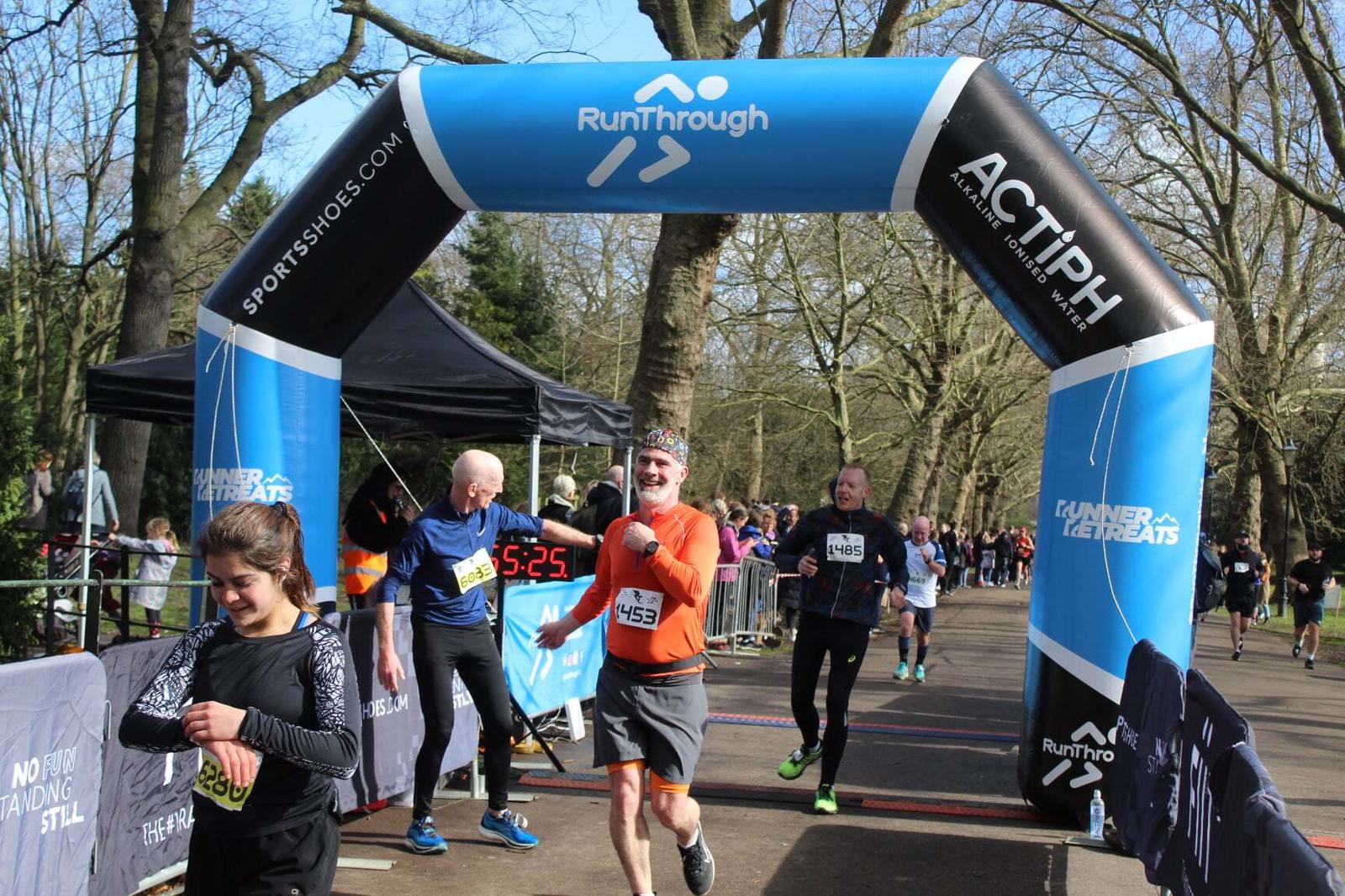 Runners cross the finish line under a blue arch marked "RunThrough" in a tree-lined park. One man in an orange shirt cheerfully raises his arm. A digital timer displays 55:25. Other participants and onlookers are visible.