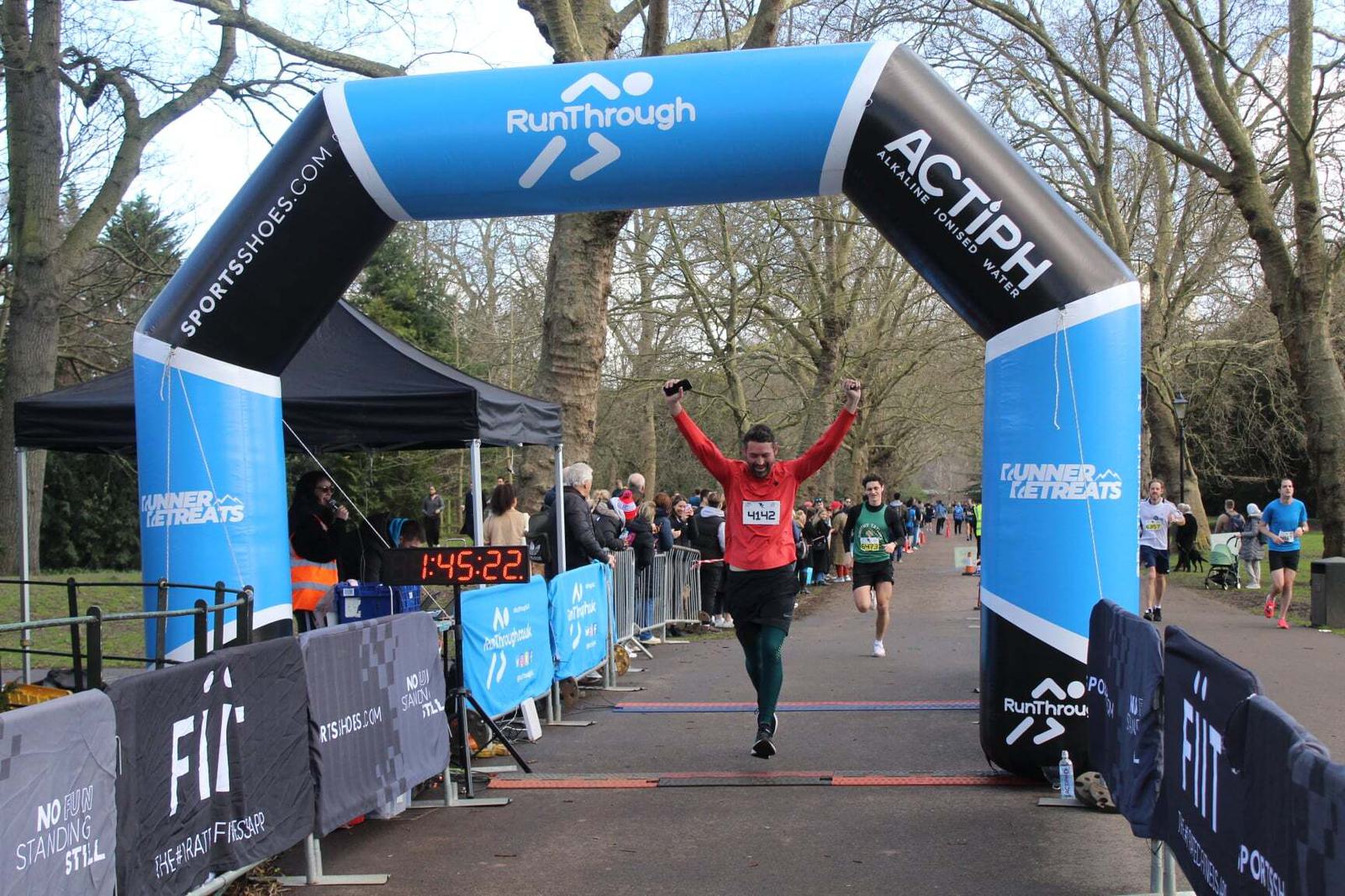 A runner crosses a blue inflatable finish line arch at a race, arms raised in victory. Other runners and spectators are visible in the background, with a clear digital timer showing 1:45:22 on the side. Trees and barriers line the pathway.