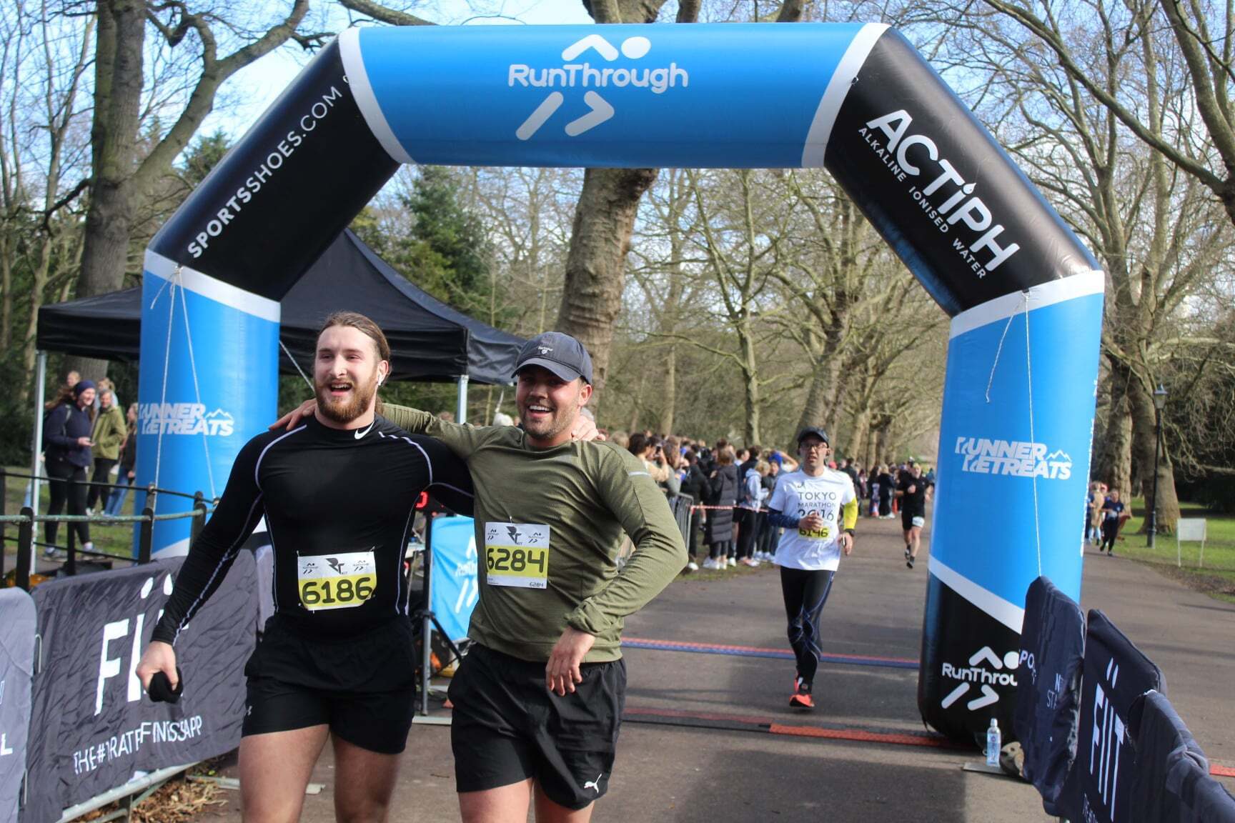Two men smile as they cross the finish line together at a running event. They are wearing race bibs numbered 6186 and 6284. A crowd and trees are visible in the background, with a blue inflatable arch overhead.