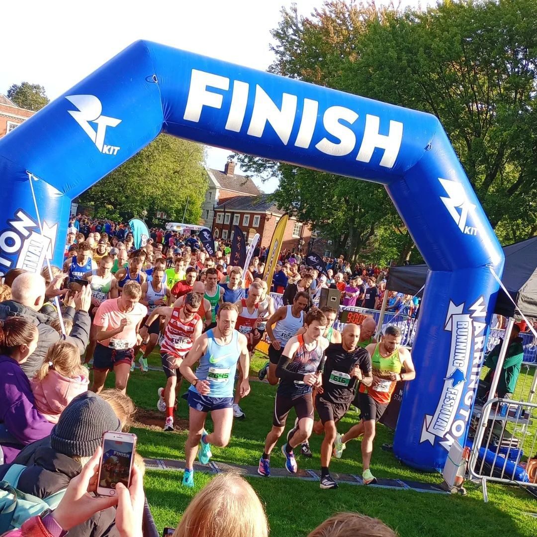 A large group of runners crosses the finish line under a blue inflatable arch that reads "FINISH." Spectators, some taking photos, stand on either side. The scene is set in a park with green grass and trees in the background.