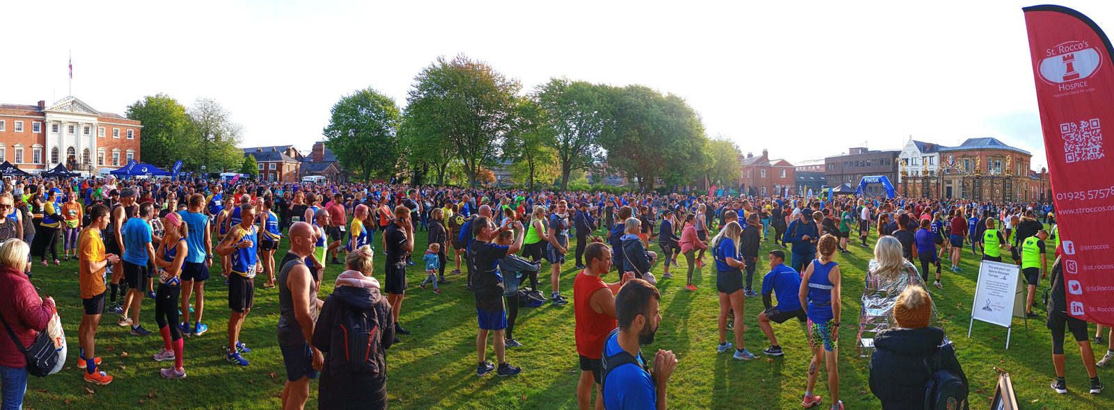 A large crowd of people gathered in an open field, many wearing running attire and bibs. Some are stretching or talking. In the background are several historic brick buildings and trees. A red banner with text and logos is visible on the right side of the image.