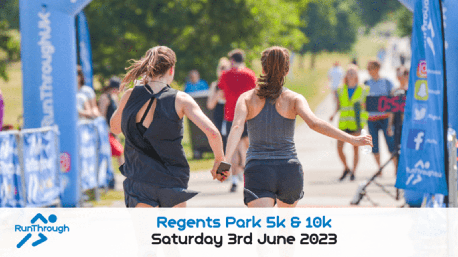 Two people hold hands as they near the finish line at the Regents Park 5k & 10k run on Saturday, 3rd June 2023. They are running past blue promotional banners and other people cheering them on. The weather appears to be sunny, and officials can be seen in the background.