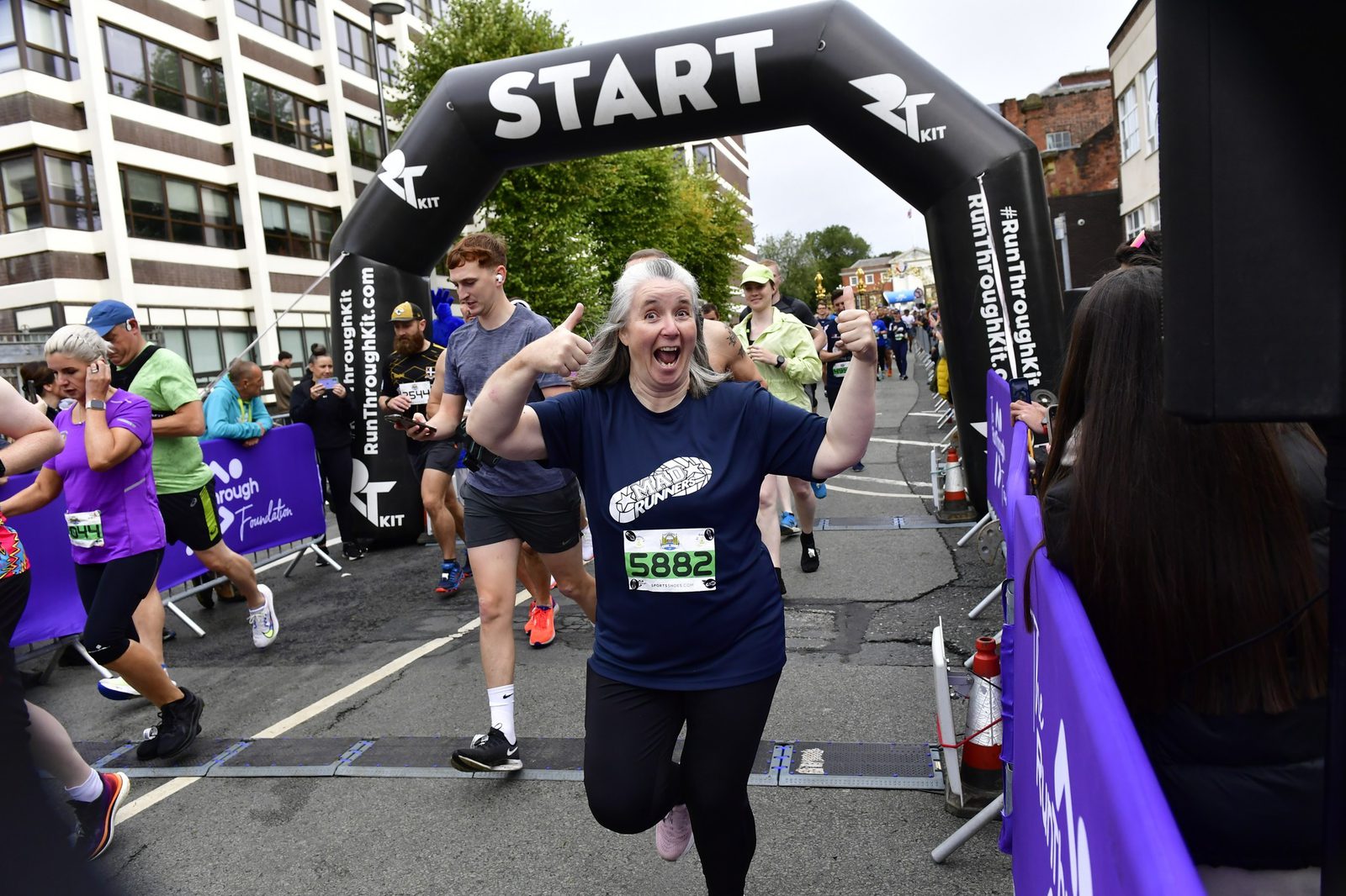 A woman with a race bib numbered 5882 excitedly poses and points at herself while passing under a black "START" arch during a running event. Other participants and spectators are visible in the background.