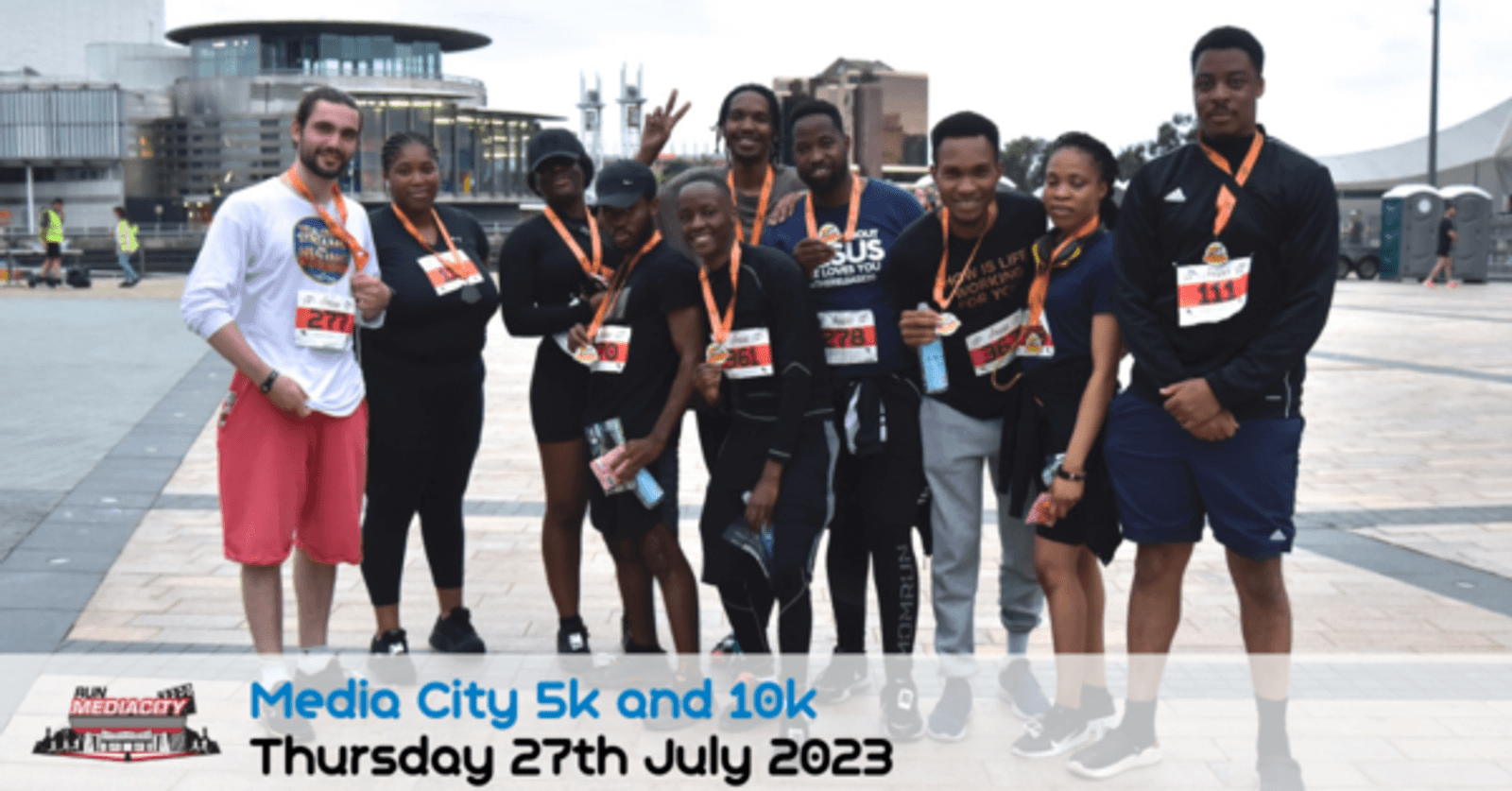 A group of smiling runners with medals around their necks pose together outdoors in Media City. They are dressed in athletic wear and standing on a paved area. A banner at the bottom reads, "Media City 5k and 10k, Thursday 27th July 2023.