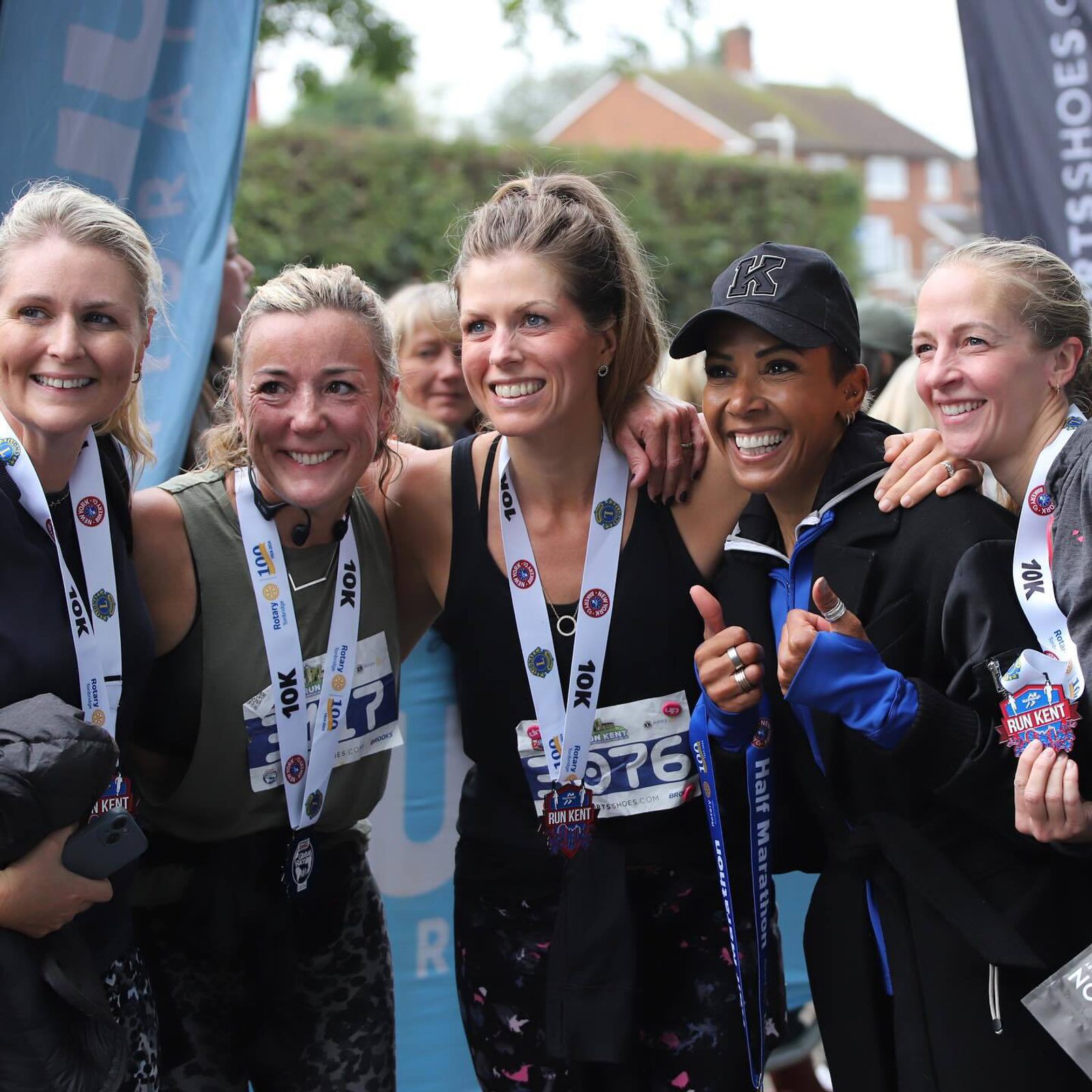 Five women in athletic gear stand close together, smiling and holding medals around their necks. They appear to be celebrating after completing a race. Trees and a glimpse of a building are in the background.