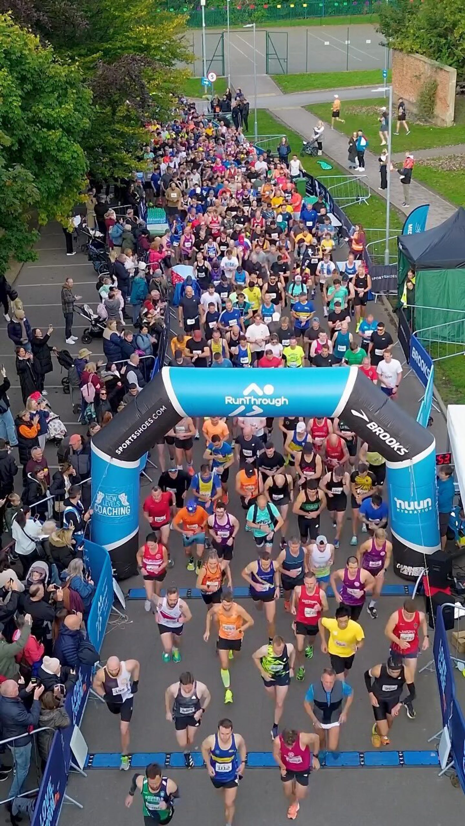 Aerial view of a large group of runners at the start of a race. They are running under a blue and white archway labeled "RunThrough." Spectators line the sides of the street, and trees and race banners are visible.