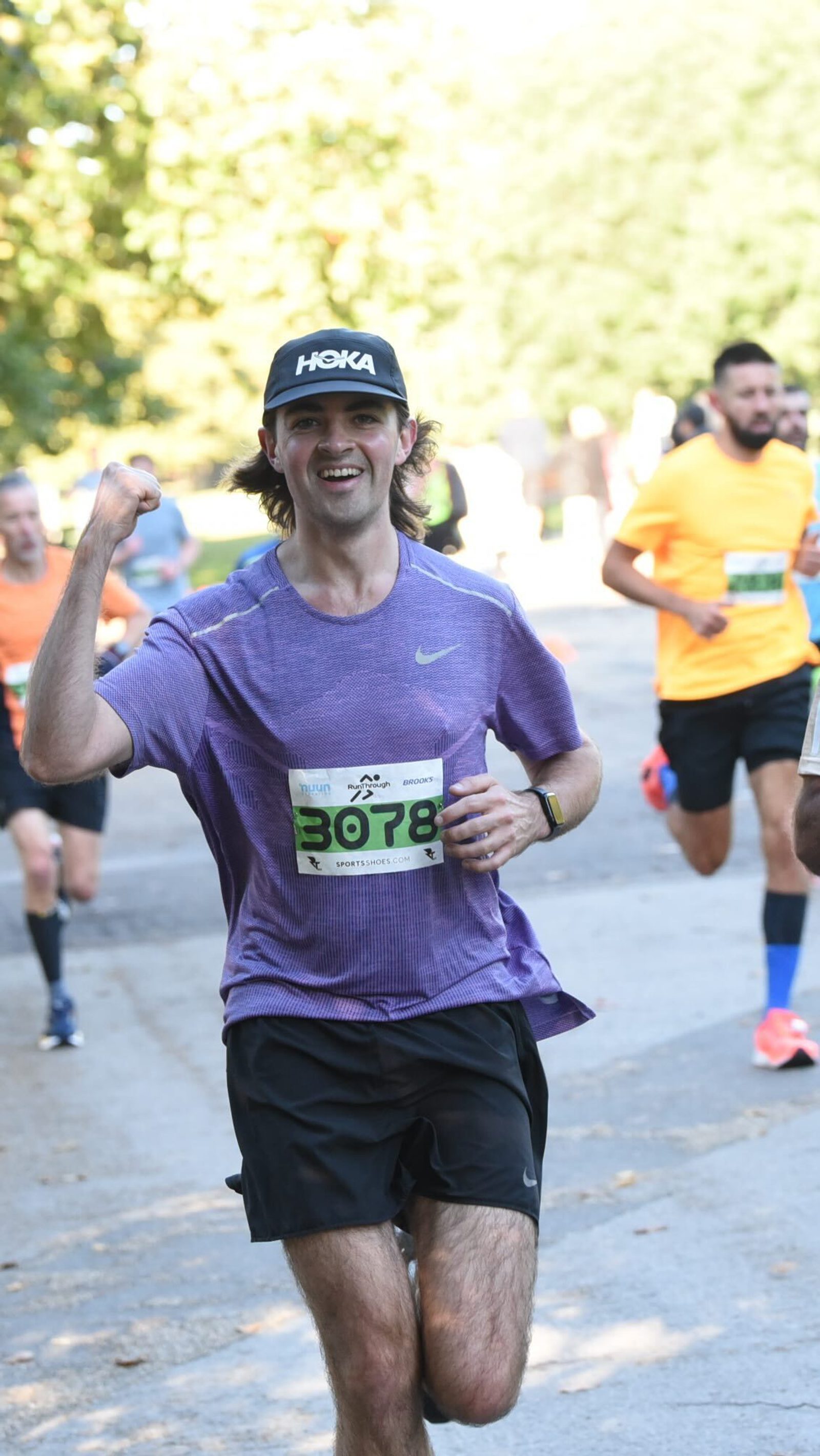A runner wearing a purple shirt and black shorts raises a fist in triumph as he runs a race. He has a race bib numbered 3078 and wears a black cap. Other runners in bright clothing are visible in the background on a sunny day.