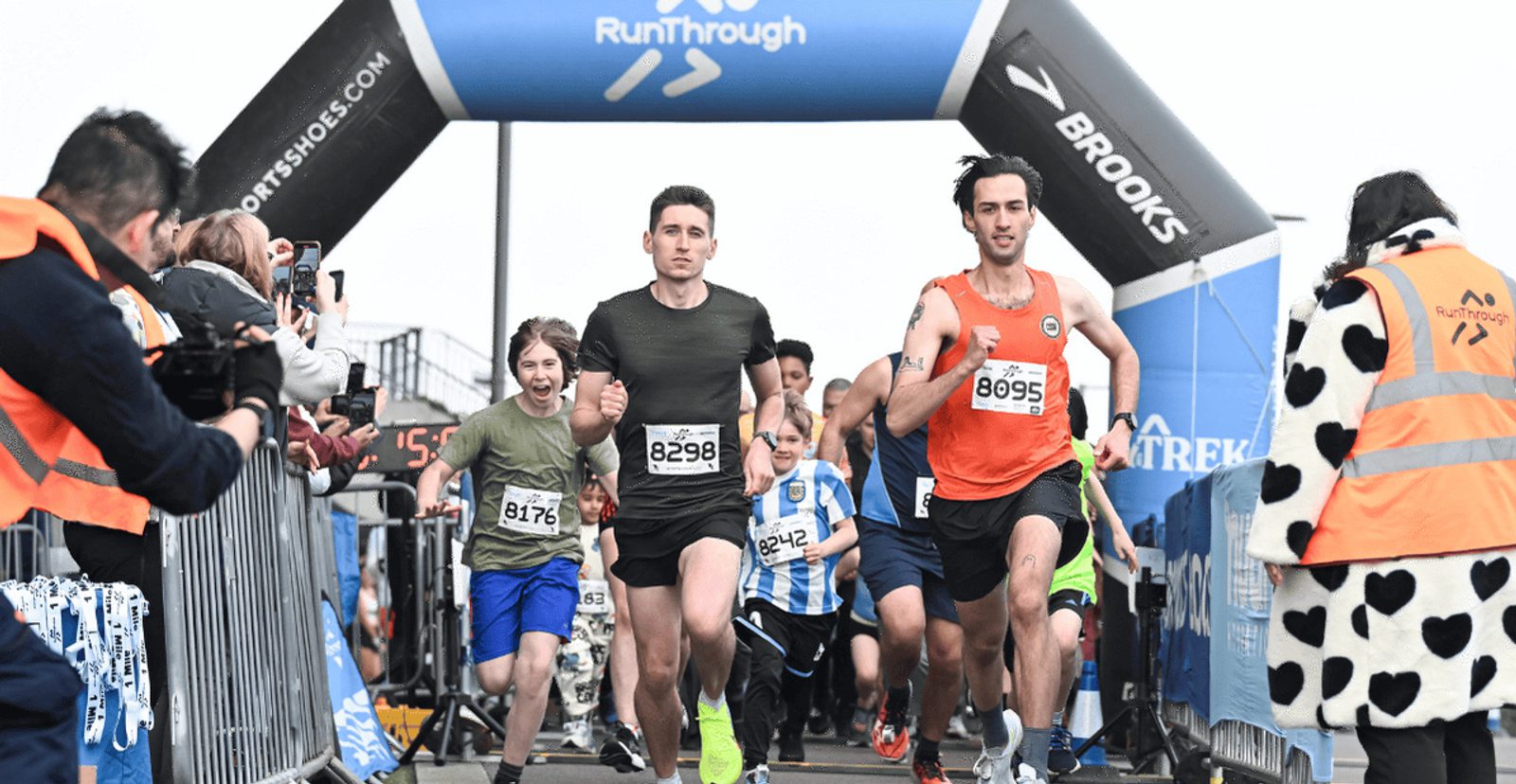 A group of runners, including a boy and two men in front, start a race under a RunThrough archway while spectators cheer and take photos beside the track.