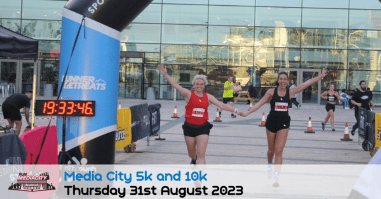 Two women are holding hands and smiling as they cross the finish line together during the Media City 5k and 10k run on Thursday, 31st August 2023. The digital clock displays 19:33:46. An archway and a glass building can be seen in the background.