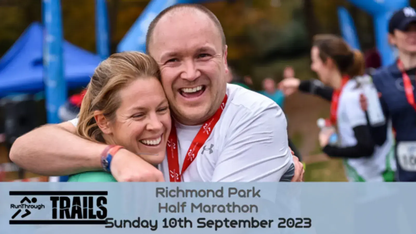 A man and woman, both wearing medals, joyfully embrace near the finish line of the Richmond Park Half Marathon. The event details and date, "Sunday 10th September 2023," are displayed at the bottom alongside the RunThrough Trails logo.