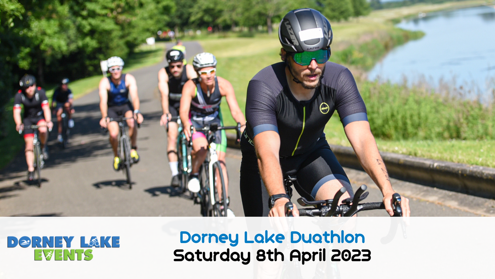 Cyclists riding on a paved path beside a lake during the Dorney Lake Duathlon. The event details, "Dorney Lake Duathlon, Saturday 8th April 2023," are displayed at the bottom in white text on a blue backdrop. The surrounding area is lush and green.