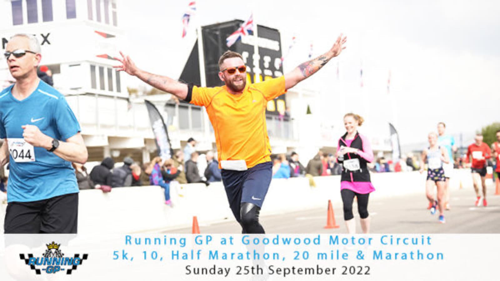 A man in a bright orange shirt and sunglasses enthusiastically spreads his arms while running on a race track during the Running GP at Goodwood Motor Circuit. Other participants are also visible, along with race banners and the event details on the image.
