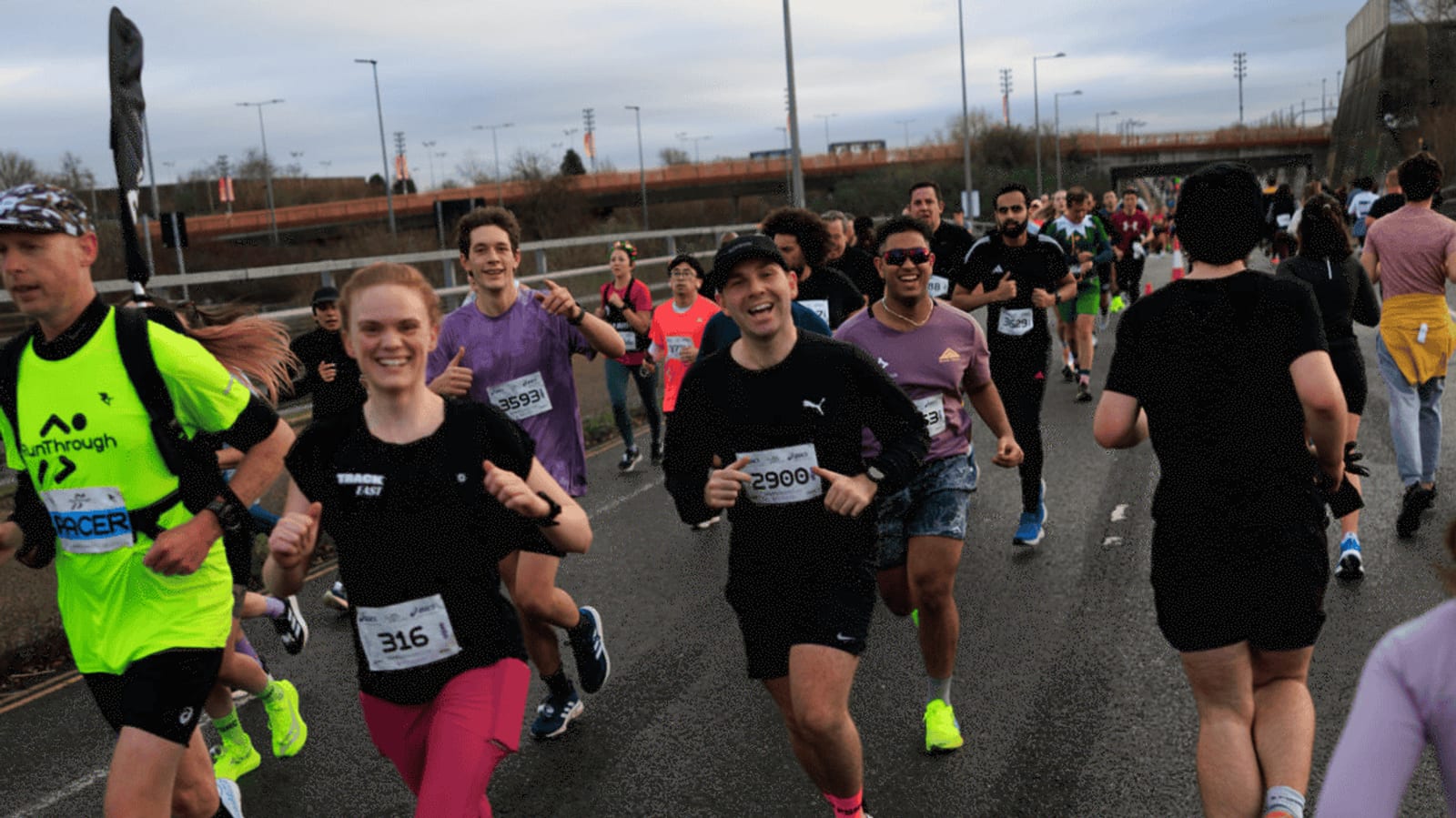 A group of runners, smiling and jogging on a road during a race, with race bibs visible. The weather is cloudy, and more participants are seen in the background, some wearing bright athletic clothing.
