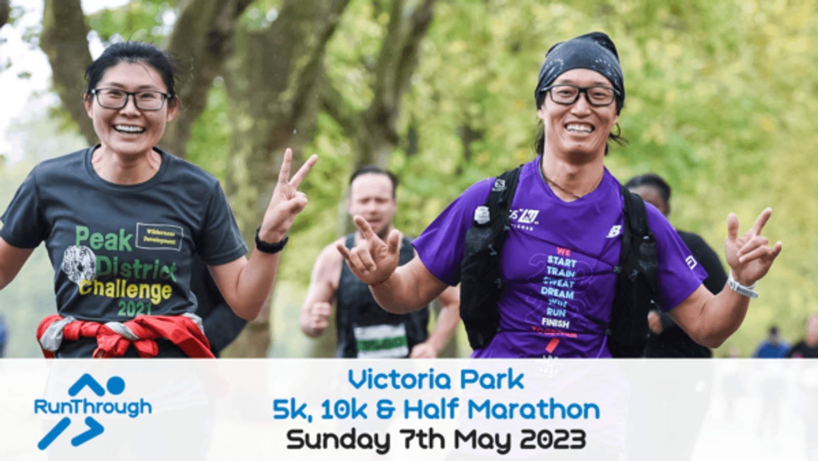 Two smiling runners are jogging outdoors during the Victoria Park event on Sunday, 7th May 2023. They are wearing glasses and race tags, with the runner on the left wearing a "Peak District Challenge" t-shirt and the one on the right in a purple shirt.