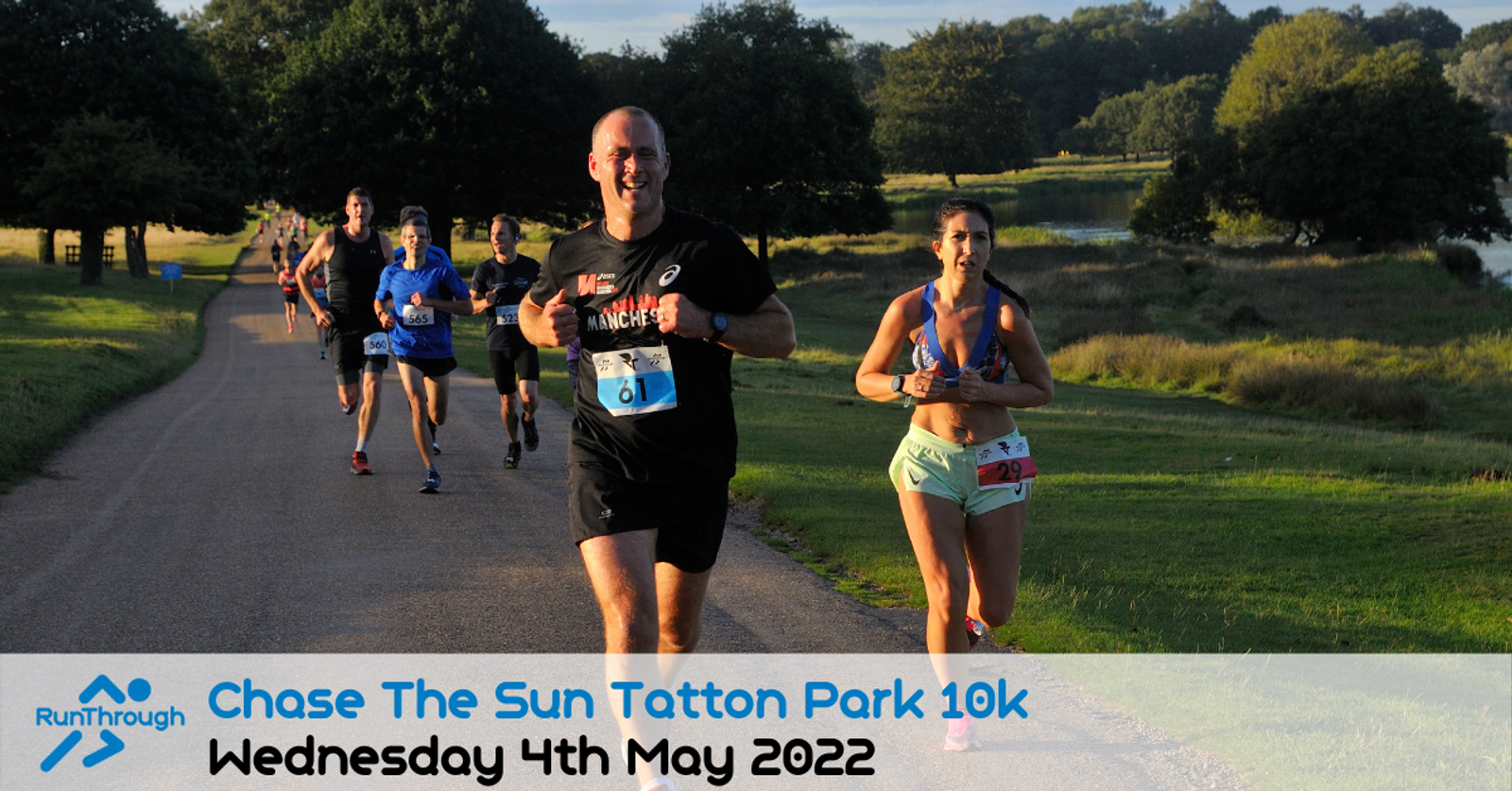 A group of runners jog along a sunny park path near trees and a pond during the "Chase The Sun Tatton Park 10k" event, held on Wednesday 4th May 2022, as shown by the event banner at the bottom of the image.