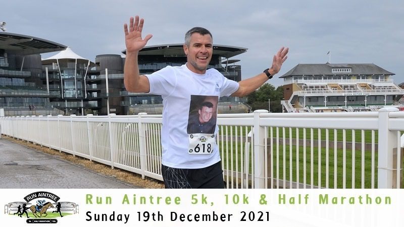 A man, wearing a white T-shirt and gray shorts with race number 618, joyfully raises his hands while running on a paved track. The event banner mentions "Run Aintree 5k, 10k & Half Marathon" taking place on Sunday, 19th December 2021. Large buildings are visible in the background.