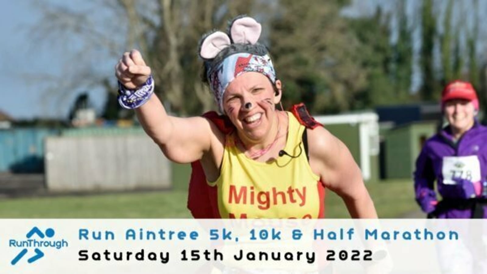 A smiling runner wearing a yellow shirt and novelty mouse ears waves during the Run Aintree 5k, 10k & Half Marathon. Another participant is visible in the background. Text overlay at the bottom reads: "Run Aintree 5k, 10k & Half Marathon Saturday 15th January 2022.