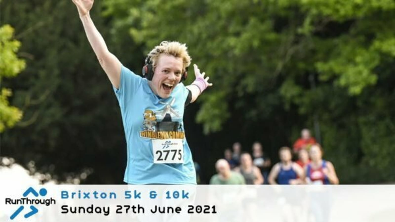 Person with headphones and race bib 2775 runs joyfully, arms spread wide. A group of runners follows behind. Trees line the background. Text reads, "Brixton 5k & 10k, Sunday 27th June 2021." Logo in the corner says "RunThrough.