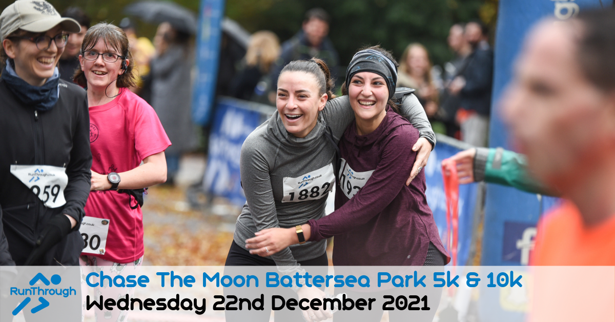 Participants smile and embrace at the finish line of the "Chase The Moon Battersea Park 5k & 10k" race held on Wednesday, December 22nd, 2021. Both wear race bibs and appear cheerful, surrounded by other participants and spectators.