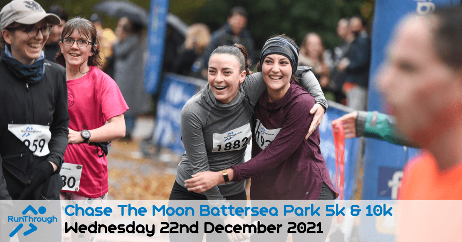 Participants smile and embrace at the finish line of the "Chase The Moon Battersea Park 5k & 10k" race held on Wednesday, December 22nd, 2021. Both wear race bibs and appear cheerful, surrounded by other participants and spectators.