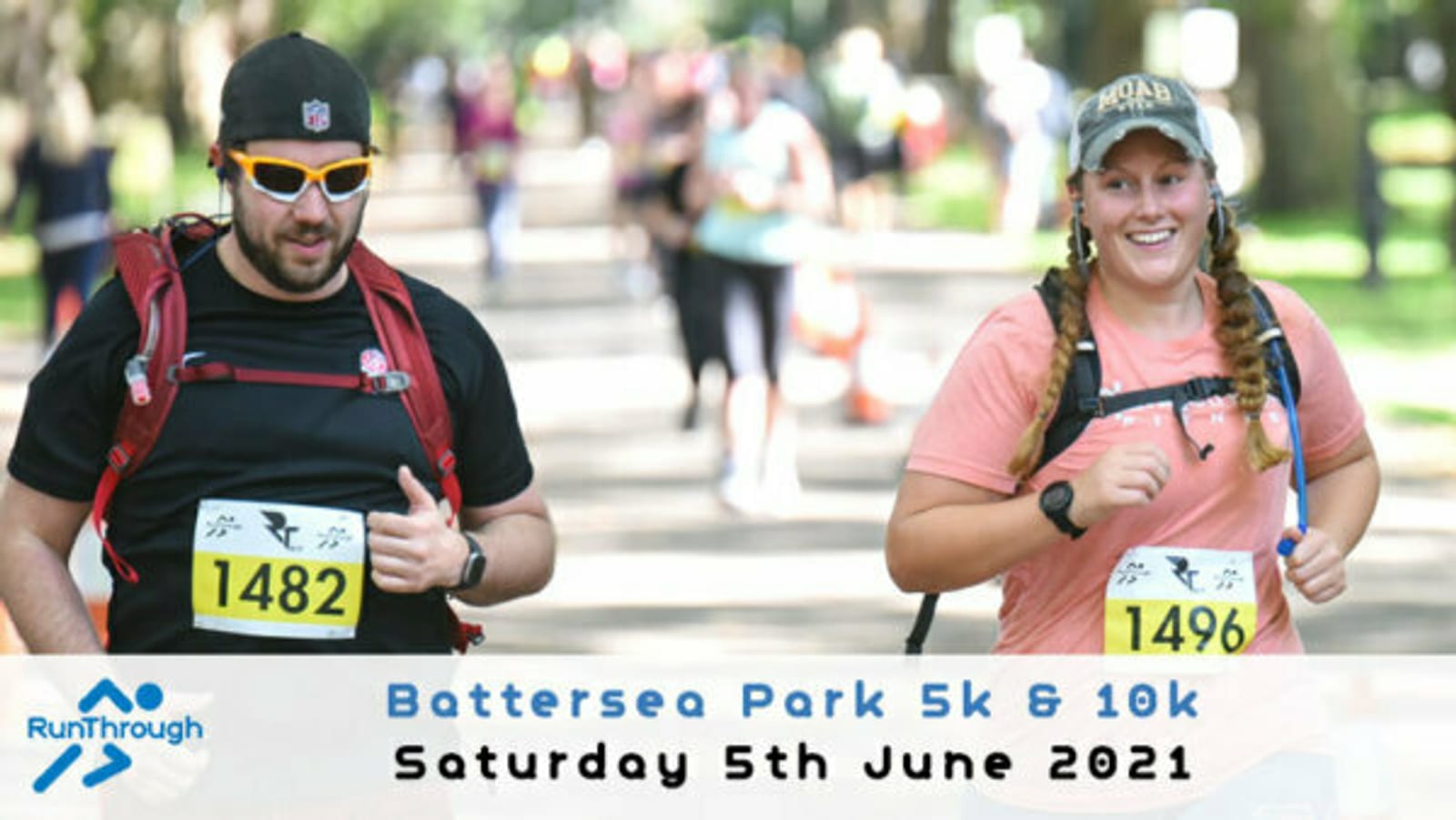 Two runners, a man and a woman, smiling and jogging in a park during a race. The man wears sunglasses, a cap, and bib number 1482, and the woman wears a cap and bib number 1496. The banner at the bottom reads "Battersea Park 5k & 10k, Saturday 5th June 2021.