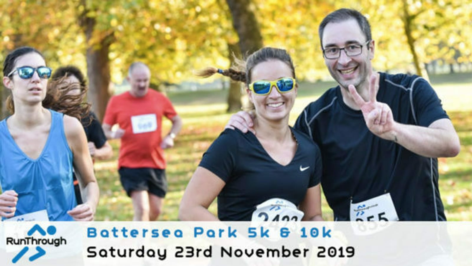 Two runners are smiling at the camera during a 5k and 10k race in Battersea Park, held on Saturday, November 23, 2019. The man on the right is holding up a peace sign. Both runners have bib numbers and are surrounded by other participants. Trees with autumn leaves are in the background.