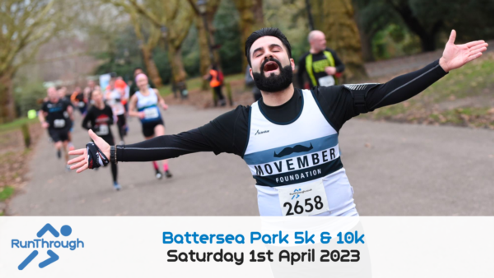 A runner with a beard and mustache wearing a Movember Foundation shirt and bib number 2658 spreads his arms wide in joy while participating in the Battersea Park 5k & 10k race on Saturday, April 1st, 2023. Other runners are visible in the background.