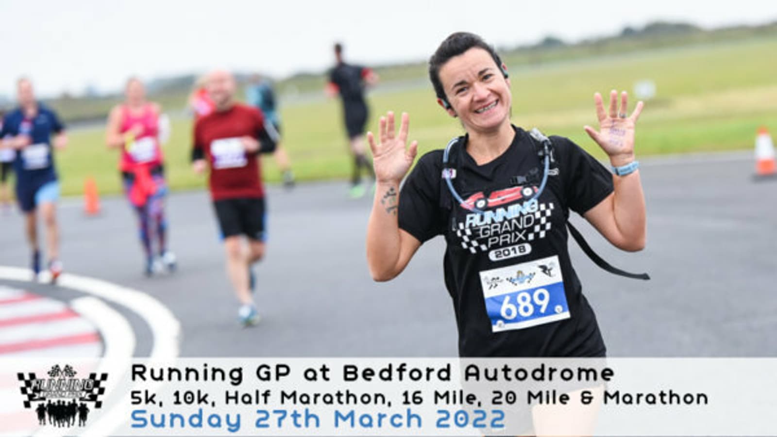 A smiling runner, wearing a black outfit and bib number 689, raises both hands in a celebratory gesture as they race on a track. Other runners are visible in the background. Text at the bottom reads "Running GP at Bedford Autodrome," and lists race events and date.