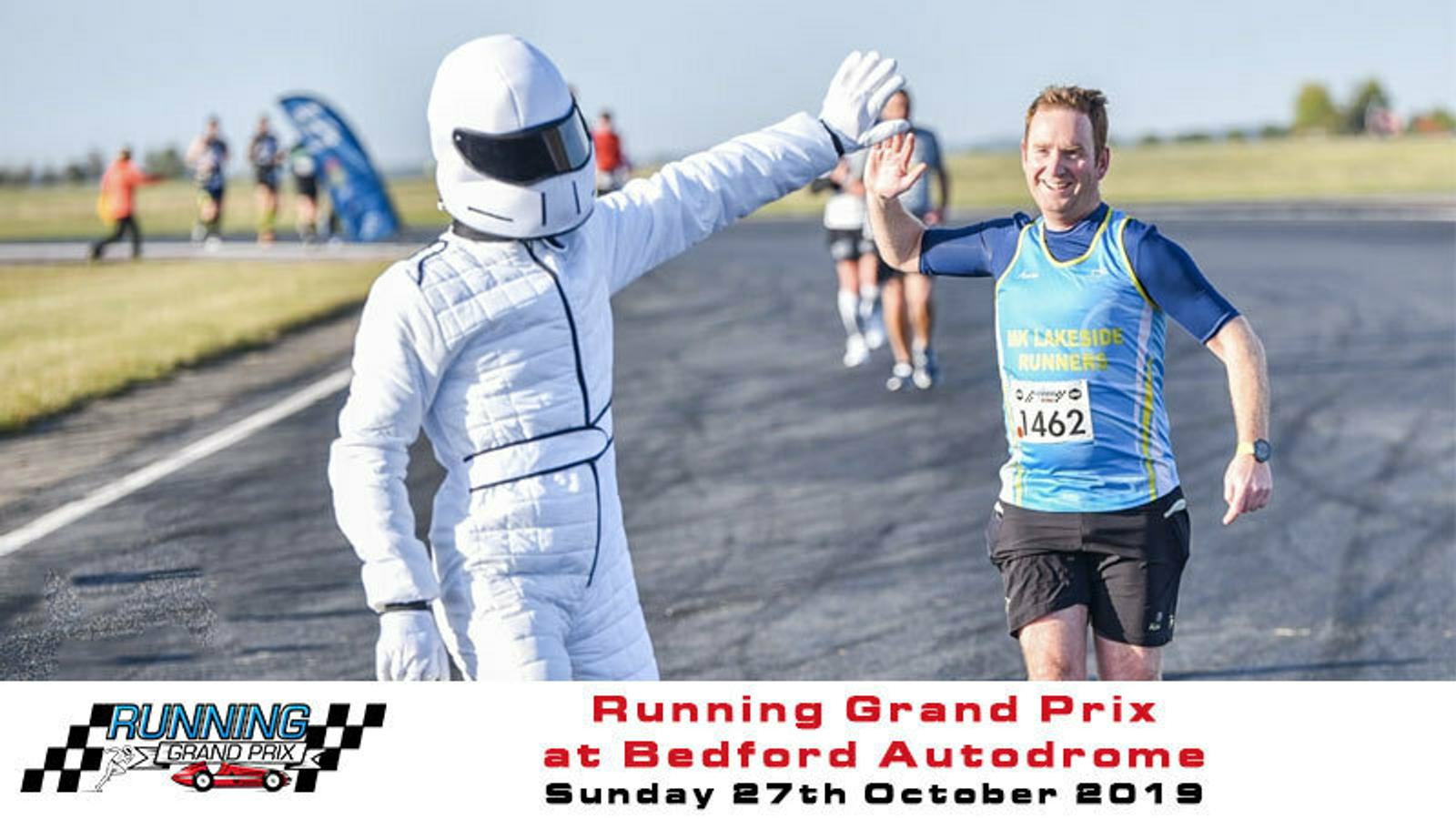 A person dressed in a white racing suit and helmet high-fives a smiling marathon runner wearing a blue and yellow running vest. In the background is a car with people and open fields. Text at the bottom reads "Running Grand Prix at Bedford Autodrome Sunday 27th October 2019.