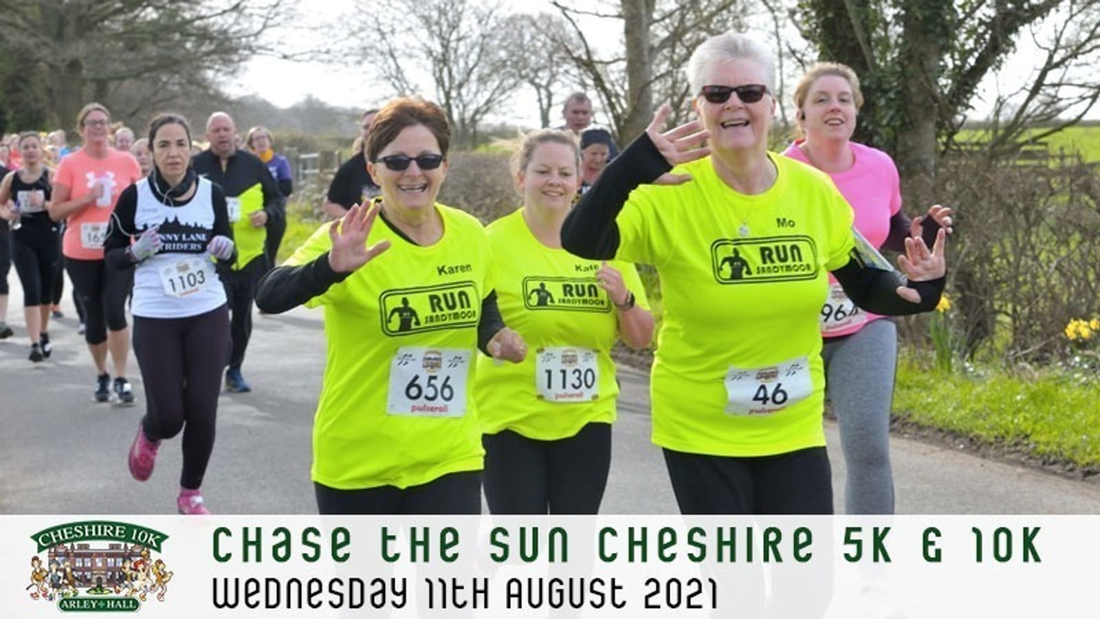 A group of runners, including two women wearing bright yellow "Run Sandbach" shirts, smile and make gestures during the "Chase the Sun Cheshire 5K & 10K" race on a rural road. The event took place on Wednesday, 11th August 2021.