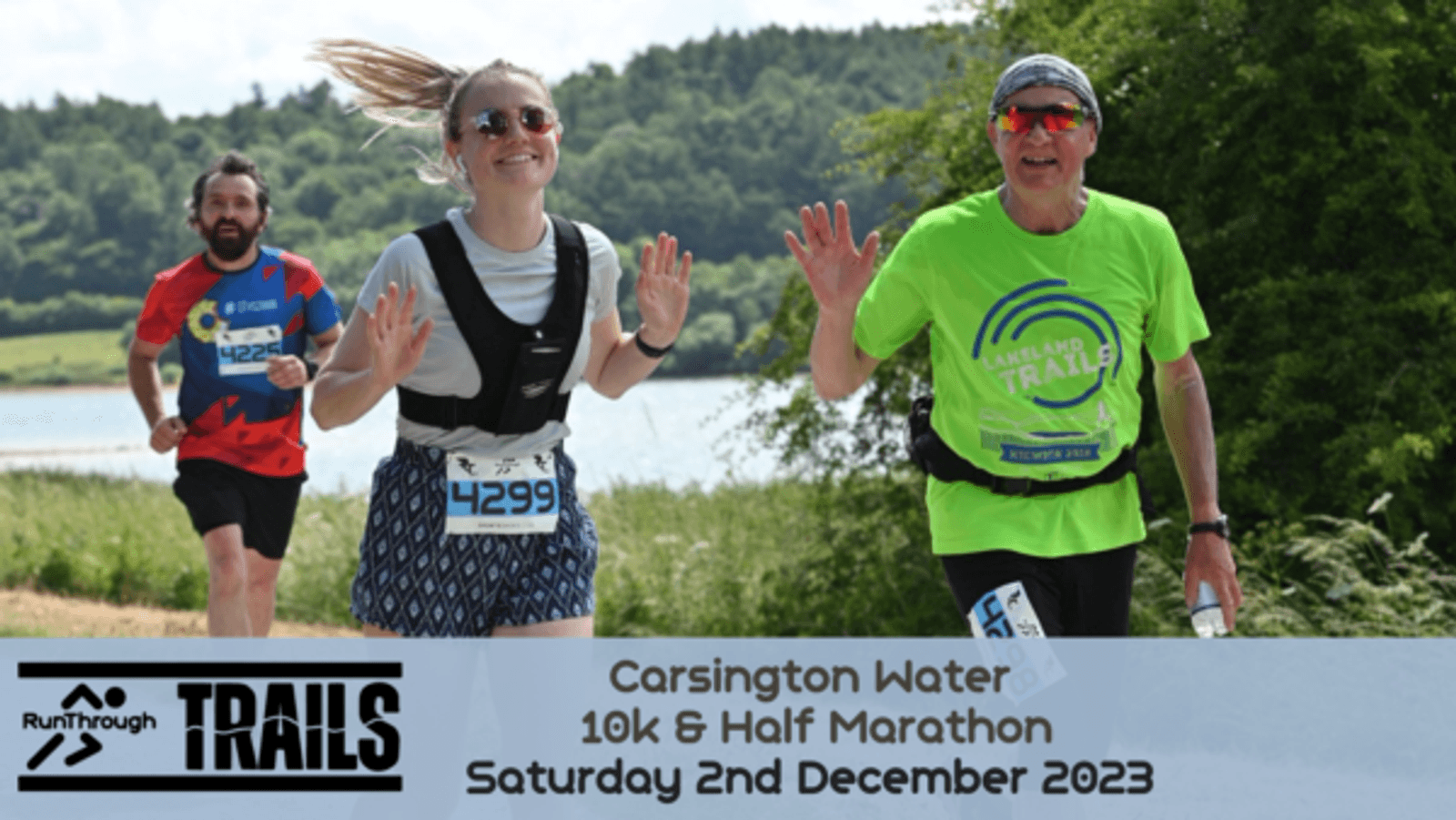 Three runners are jogging outdoors during the Carsington Water 10k & Half Marathon event. They are smiling and waving at the camera under a clear sky with trees and water in the background. The event details and date are displayed at the bottom of the image.