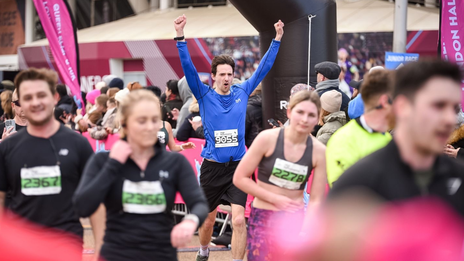 A group of people are running in a race, wearing bibs with numbers. There is a man in a blue shirt with the number 9055 in the background, raising his arms in celebration. Other runners and spectators are blurred in the foreground.