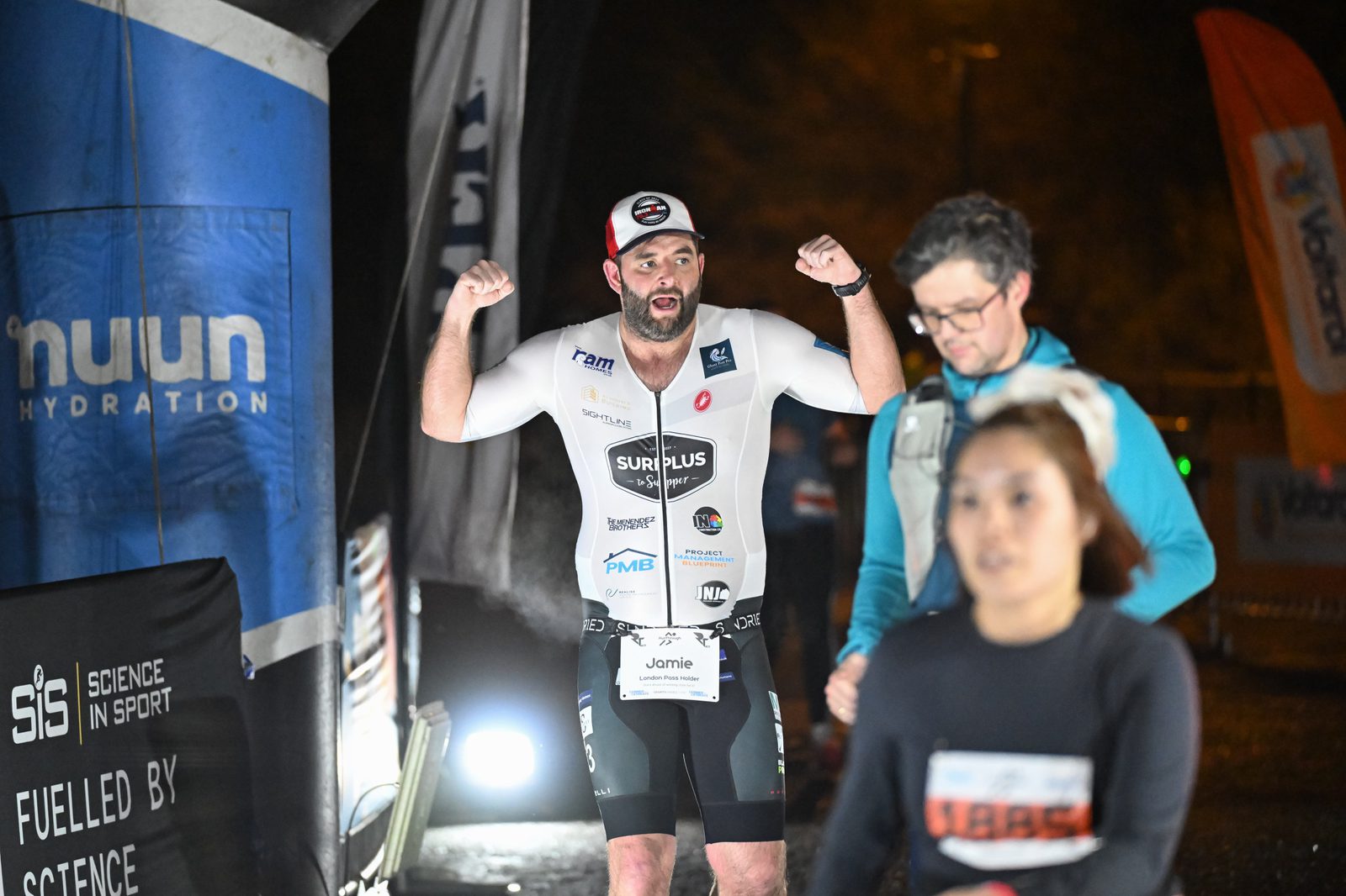 A triathlete wearing a white and black suit raises his arms in celebration at a race finish line, while two other participants walk ahead of him. The scene is lit with bright lights and branded banners are visible in the background.