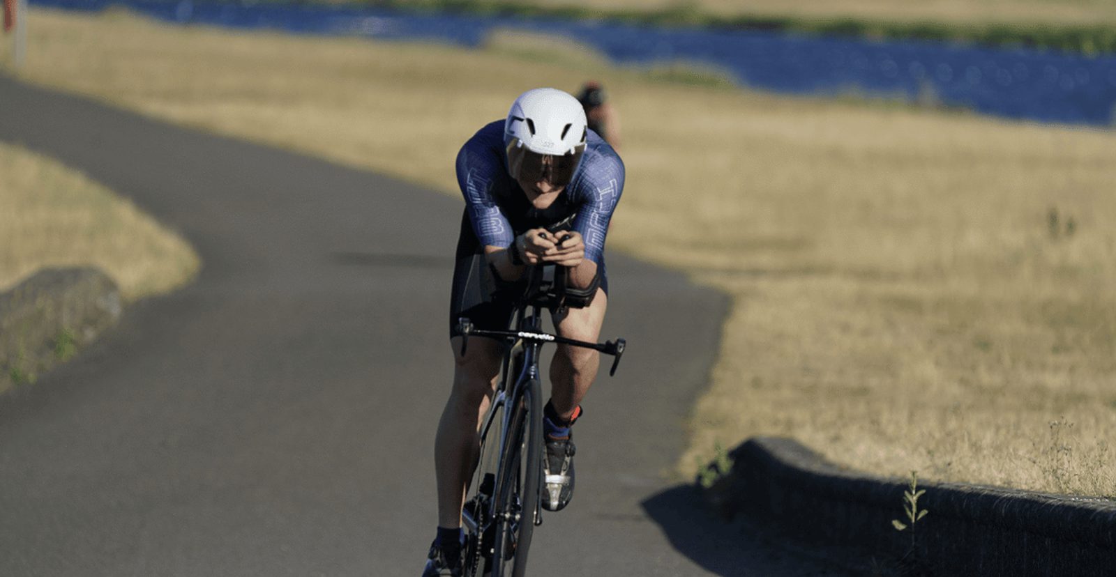 A cyclist wearing a white helmet and blue suit rides a road bike on a paved path through a dry, grassy landscape with water visible in the background.