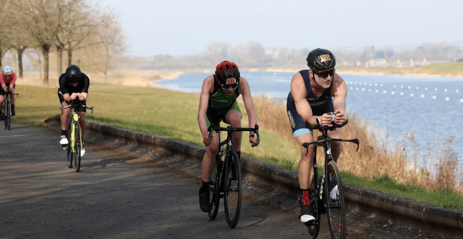 Four cyclists race along a paved road beside a river, with trees lining one side and grassy banks on the other. The riders are leaning forward intensely, wearing athletic gear and helmets.
