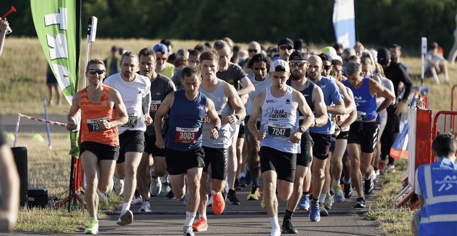 A group of runners wearing numbered bibs start a race on a paved outdoor path, surrounded by grassy fields and trees in the background. Some spectators and event staff are visible along the sides.