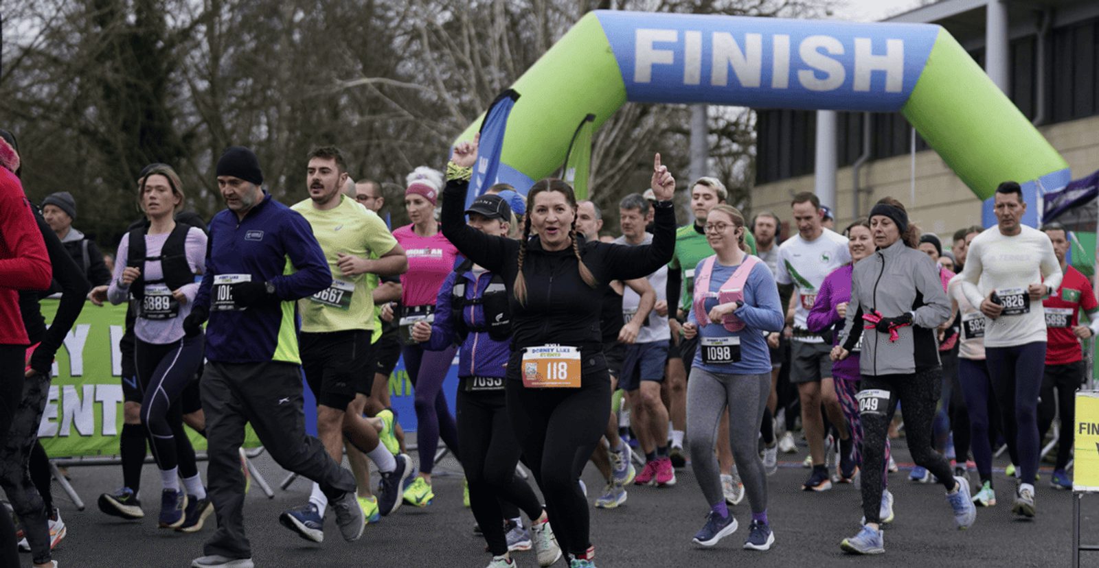 A group of runners crosses the finish line at an outdoor race event. The participants wear athletic clothing and race bibs, with trees and a large blue and green "FINISH" arch visible in the background.