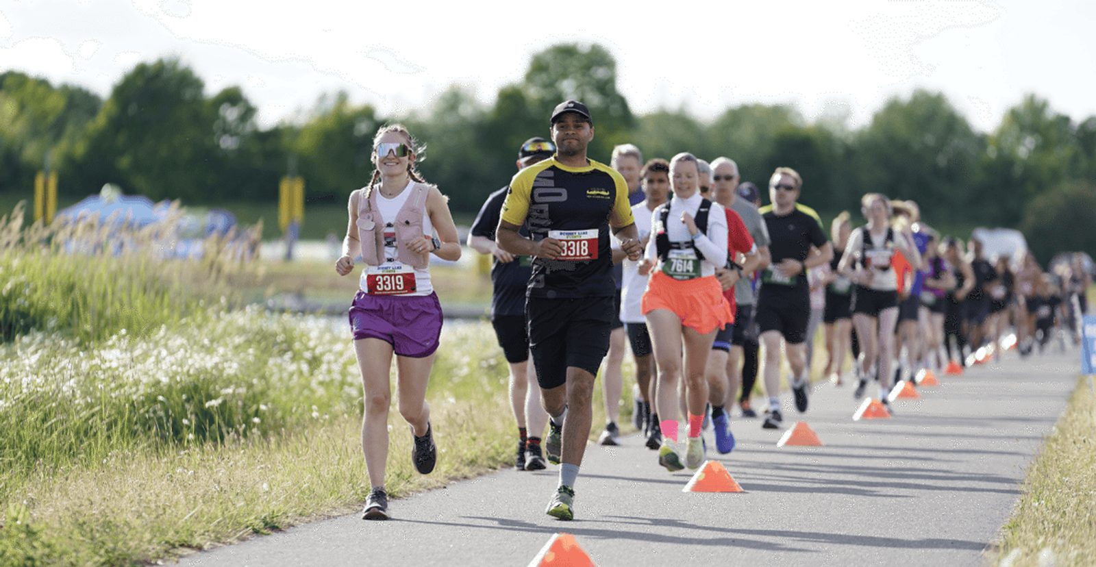 A group of runners participate in a race on a paved path lined with orange cones, surrounded by greenery and trees under a bright sky.